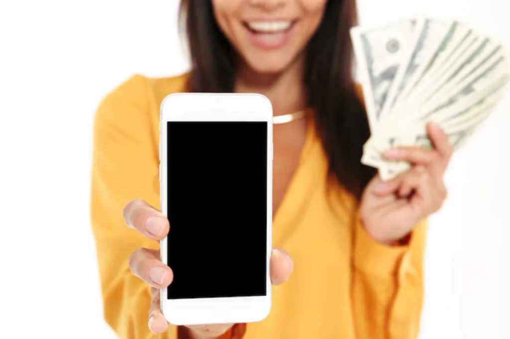 A woman in a yellow shirt smiles while holding a smartphone and a stack of cash, suggesting financial success or opportunity.