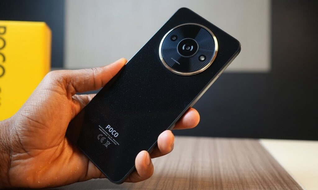 A person holding a black POCO smartphone, showcasing its circular camera lens and sleek design, with a yellow POCO box in the background.