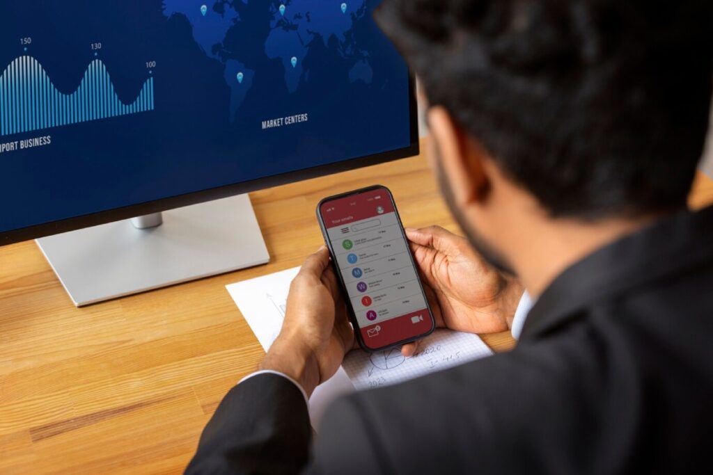 A businessman checks his smartphone displaying email notifications, with a data visualization map on a monitor in the background.