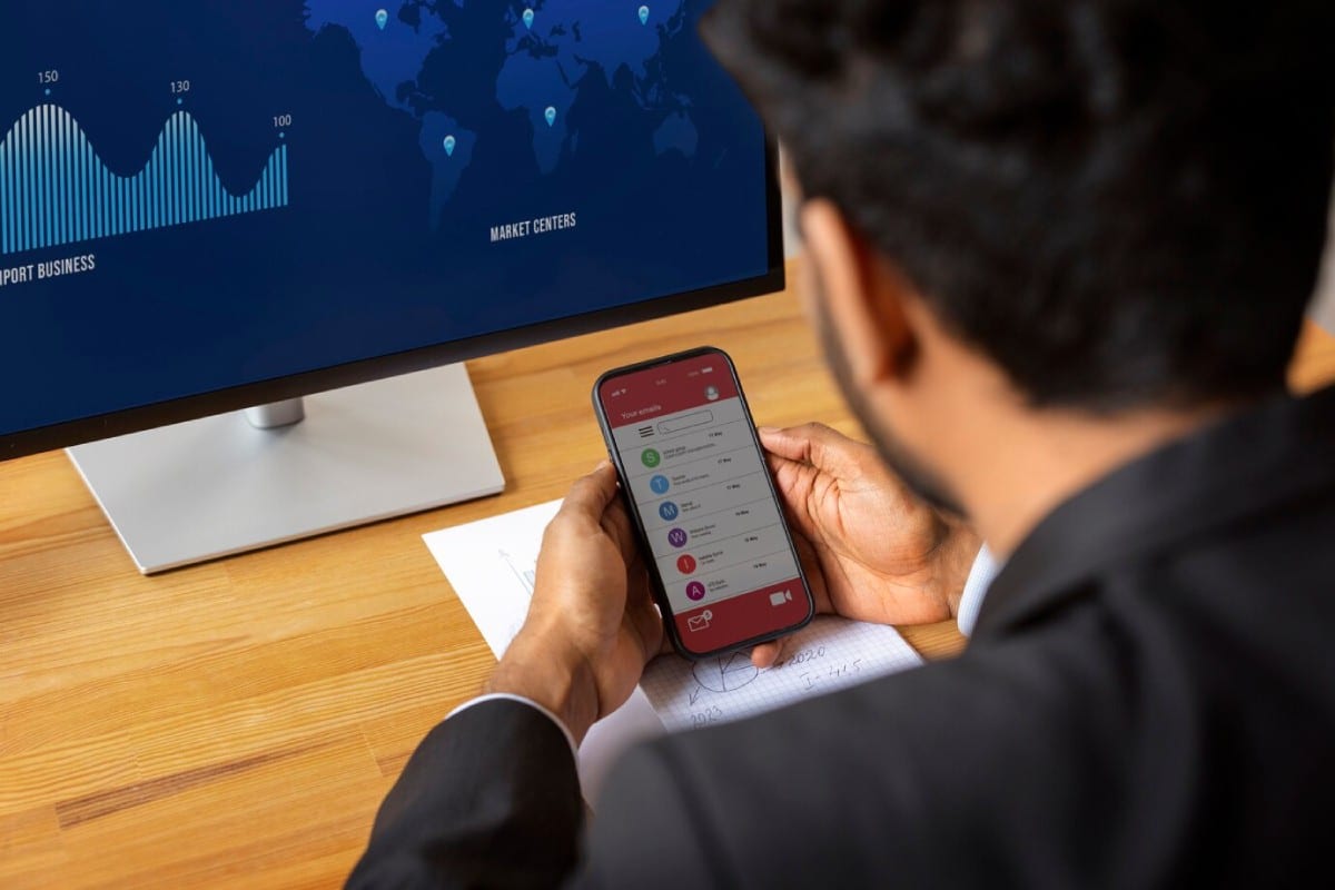 A businessman checks his smartphone displaying email notifications, with a data visualization map on a monitor in the background.