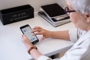 An older adult uses a smartphone on a desk, interacting with a smart home app to adjust settings, surrounded by books and decorative boxes.