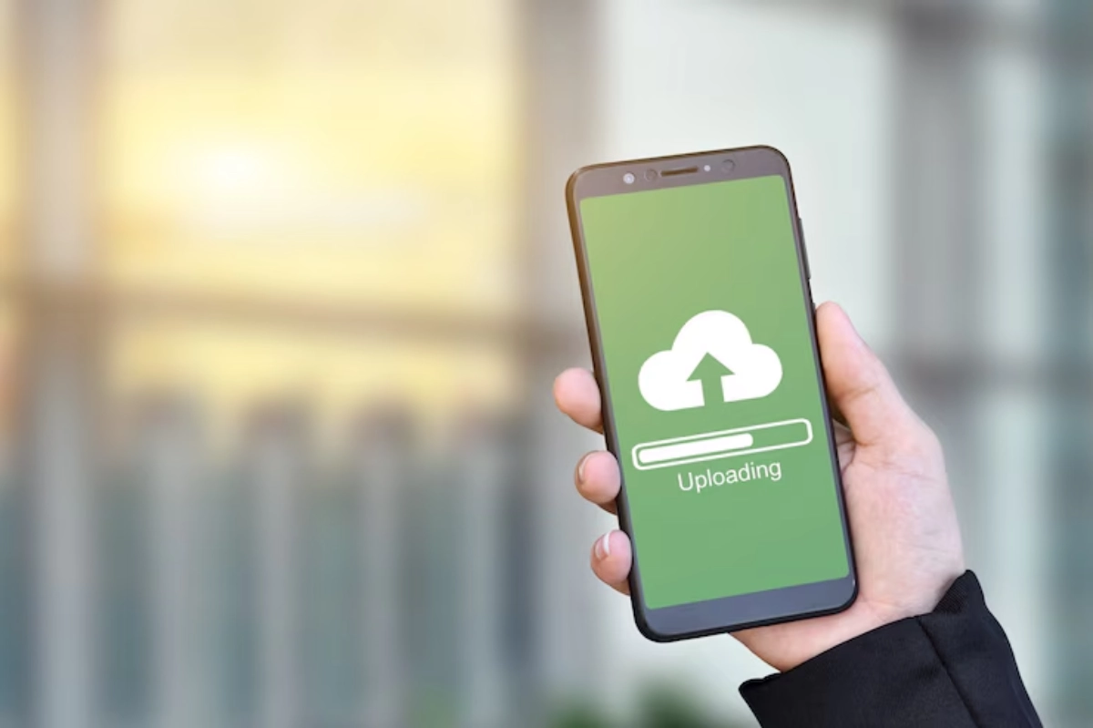 A hand holds a smartphone displaying an Uploading screen with a cloud icon and progress bar, against a blurred outdoor background.
