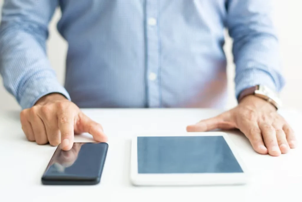 A person in a blue checkered shirt uses a smartphone with one hand while resting an arm on a tablet, both on a white surface.
