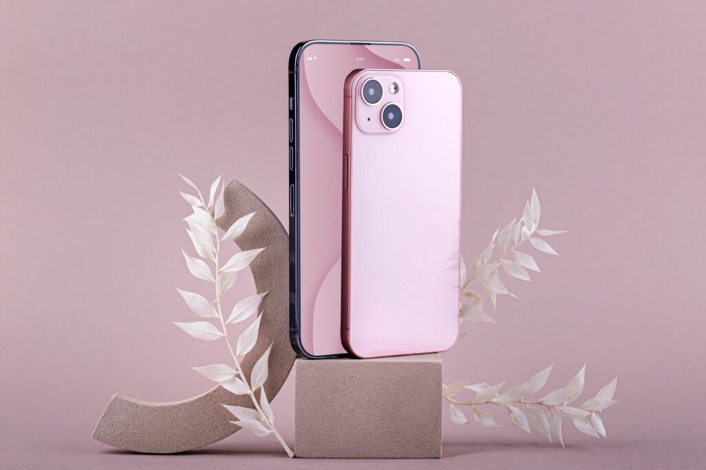 A pink smartphone stands beside a black one, elegantly displayed on a geometric pedestal with delicate white foliage against a soft pink backdrop.