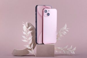A pink smartphone stands beside a black one, elegantly displayed on a geometric pedestal with delicate white foliage against a soft pink backdrop.