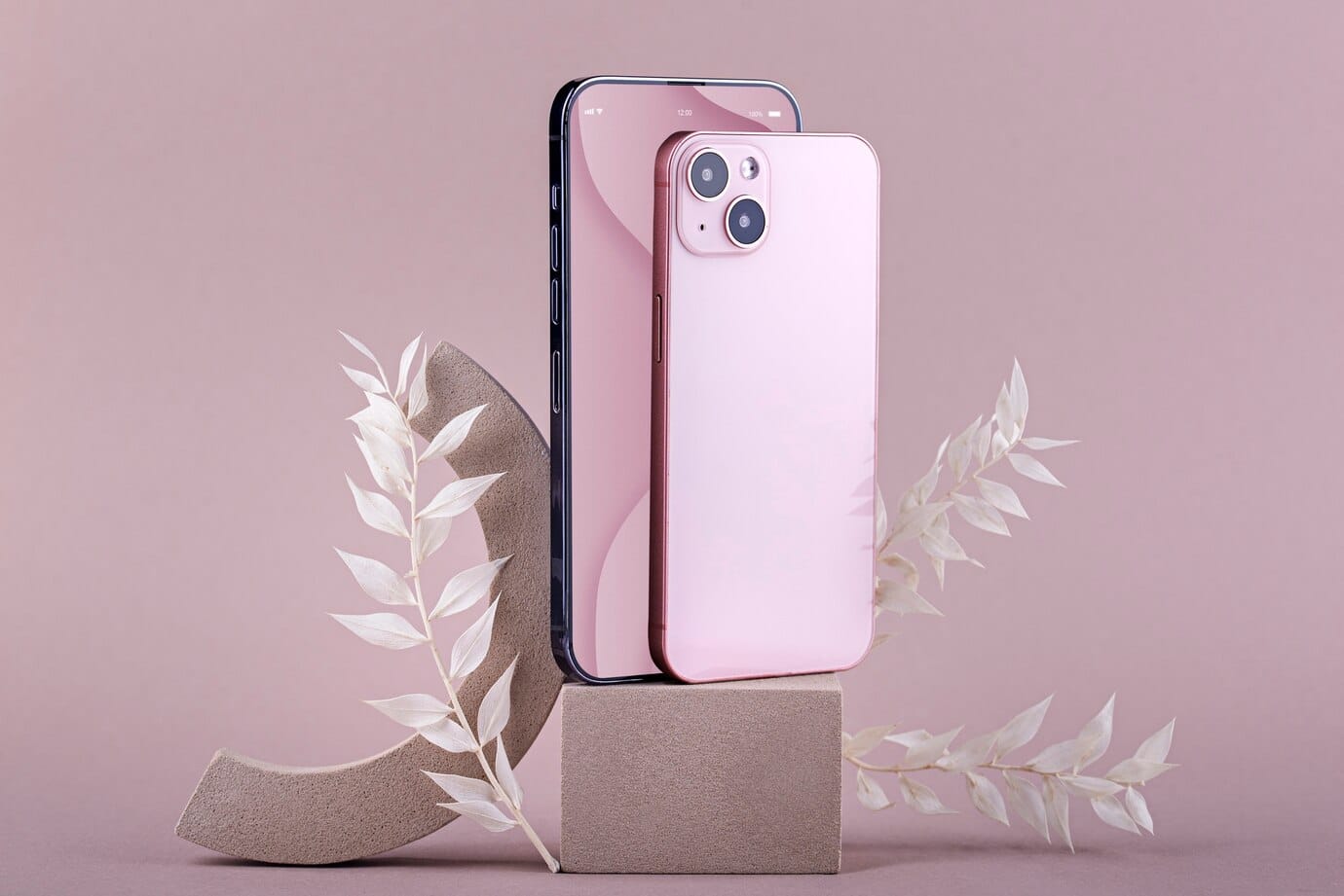 A pink smartphone stands beside a black one, elegantly displayed on a geometric pedestal with delicate white foliage against a soft pink backdrop.