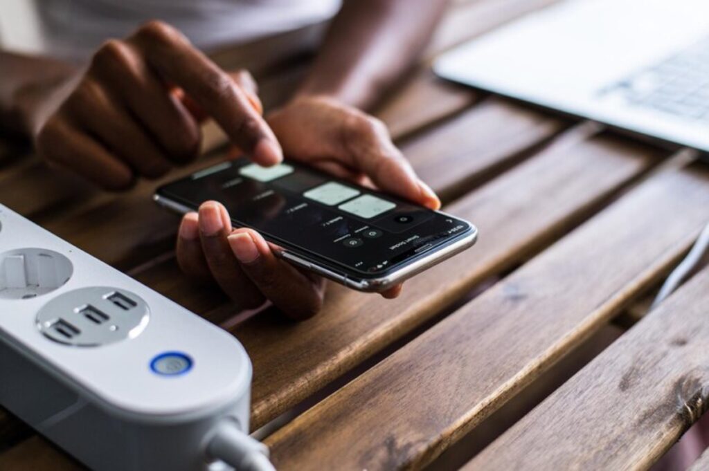 A person uses a smartphone to control devices, with a power strip and laptop visible on a wooden table.