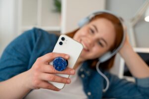 A person in a denim shirt holds a white smartphone with a blue pop socket, wearing headphones in a cozy indoor setting.