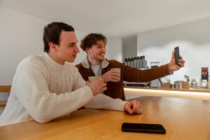 Two young men enjoy drinks while taking a selfie in a modern café, with a warm wooden table and soft lighting in the background.