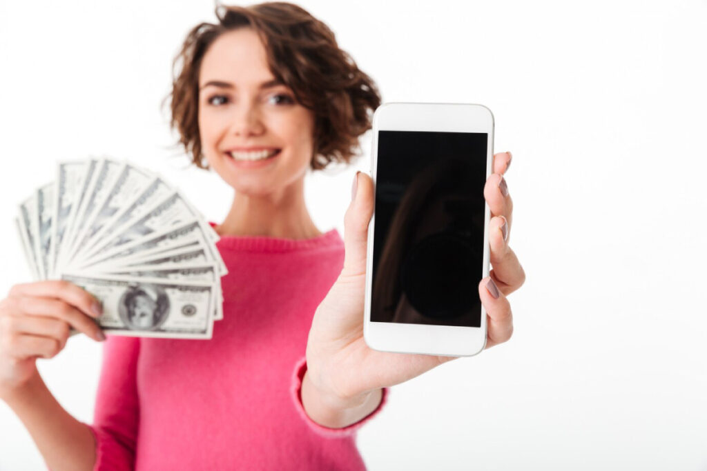 A person holds a smartphone in one hand and a fan of cash in the other, smiling against a plain white background.
