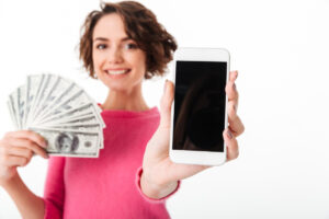 A person holds a smartphone in one hand and a fan of cash in the other, smiling against a plain white background.