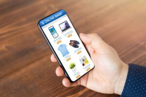 A person holds a smartphone displaying an online shopping app with various product categories, set against a wooden table background.