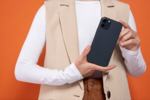 A person wearing a beige vest and white long-sleeve shirt holds a dark smartphone against a vibrant orange background.