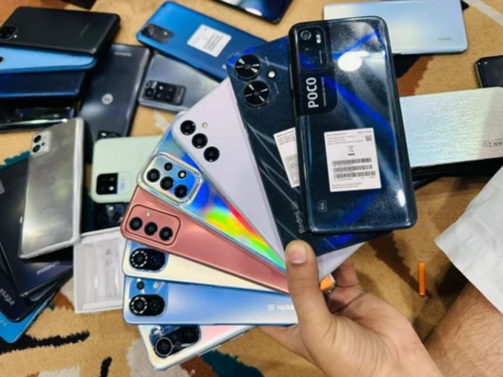 A hand holding multiple smartphone models in various colors, surrounded by other phones on a textured surface.