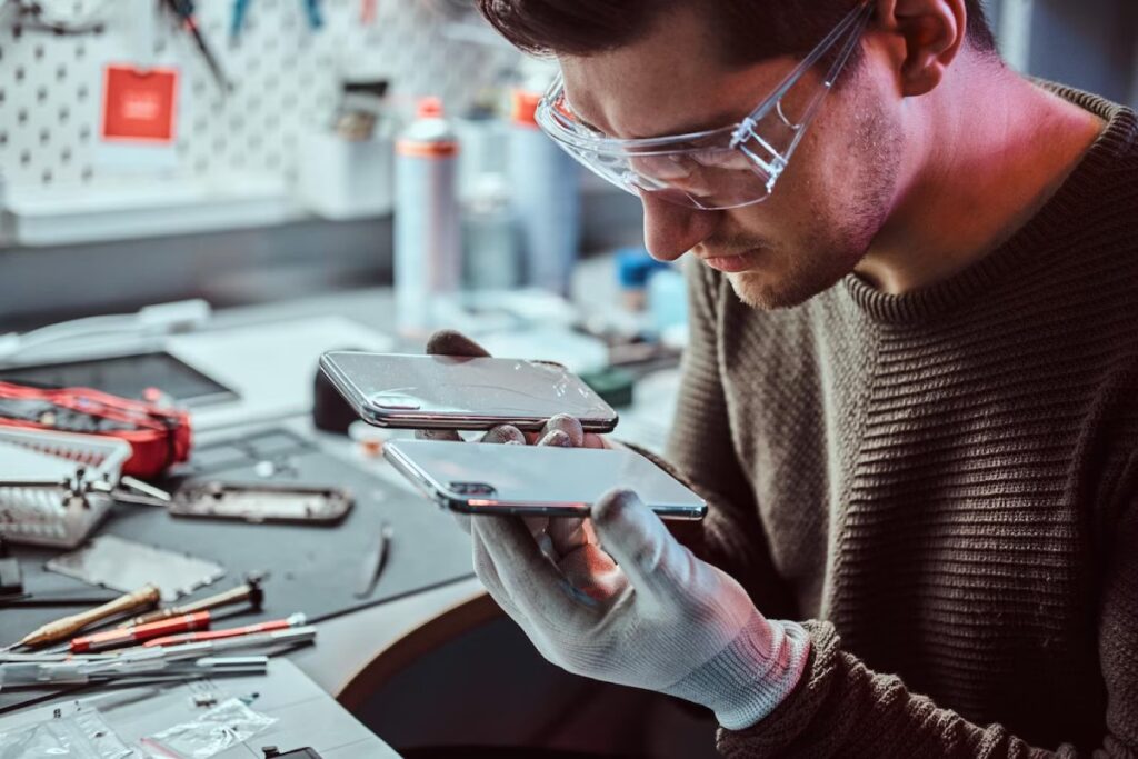 A technician in gloves inspects two smartphones, one cracked, on a cluttered workbench filled with tools and equipment.
