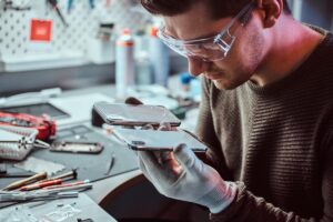 A technician in gloves inspects two smartphones, one cracked, on a cluttered workbench filled with tools and equipment.