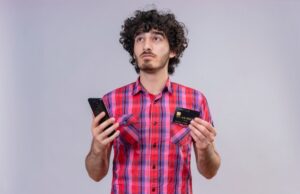 A person wearing a plaid shirt holds a smartphone in one hand and a credit card in the other, set against a gray background.