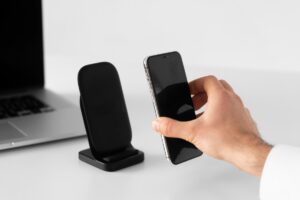 A hand positions a smartphone above a black wireless charger next to a laptop on a minimalistic white desk.