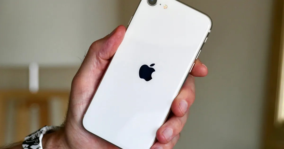 A person's hand holding a white Apple iPhone with a visible black Apple logo on the back against a blurred background.