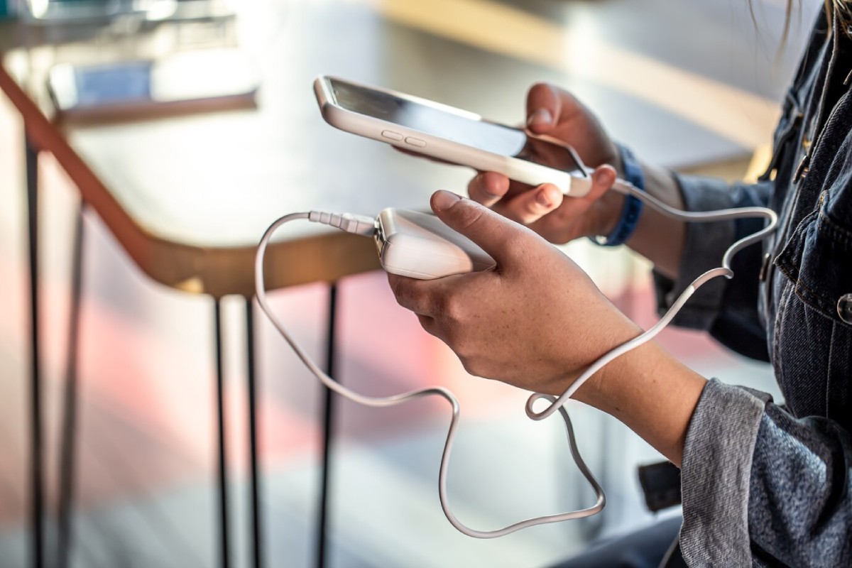 A person holds a smartphone and a portable charger, connecting them with a charging cable while seated at a table.