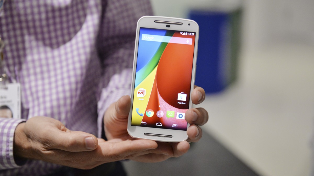A hand holds a white Motorola smartphone displaying a colorful home screen with app icons, set against a blurred background.
