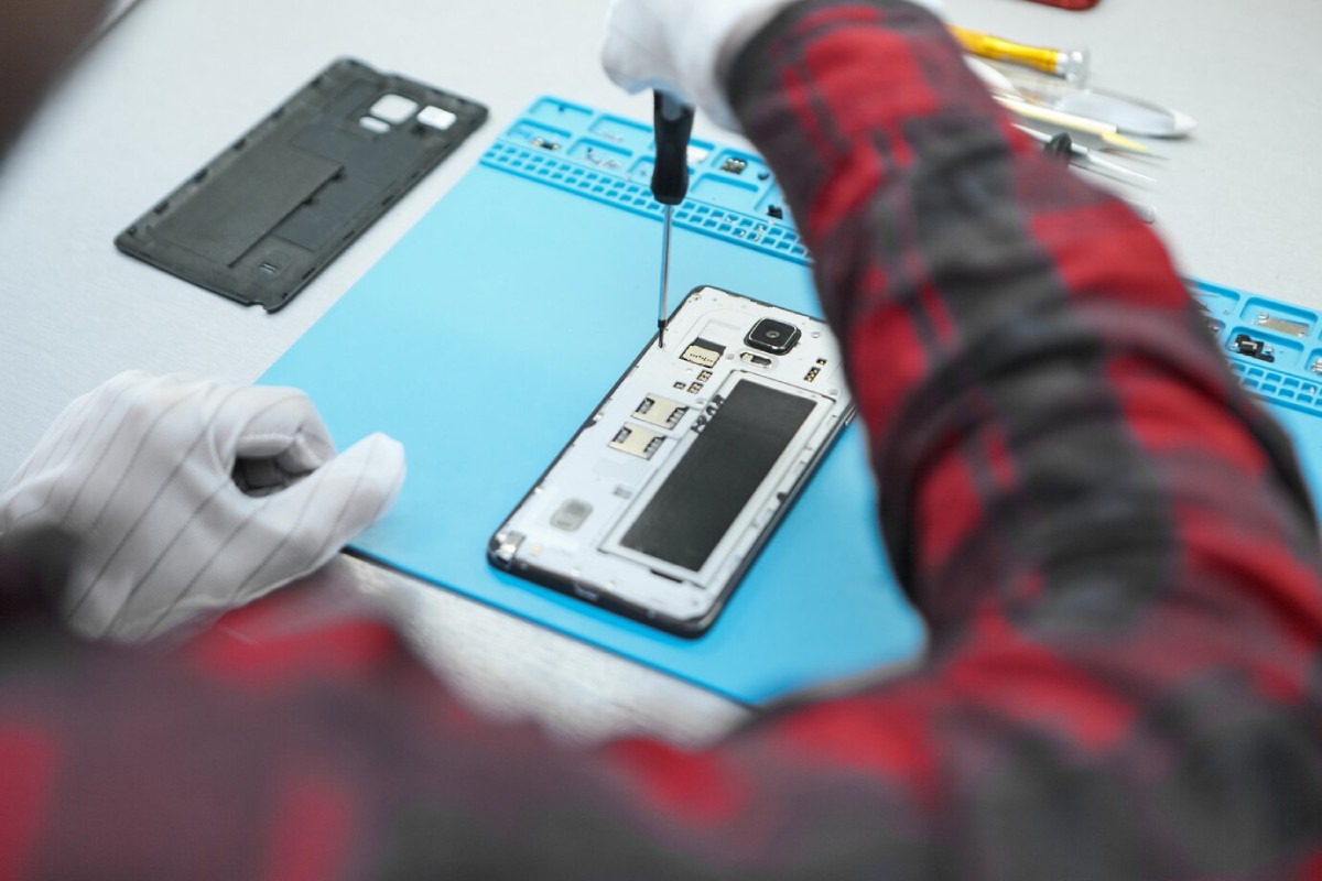 A technician in gloves uses a screwdriver to repair a smartphone, with tools and the phone's back cover nearby on a work surface.