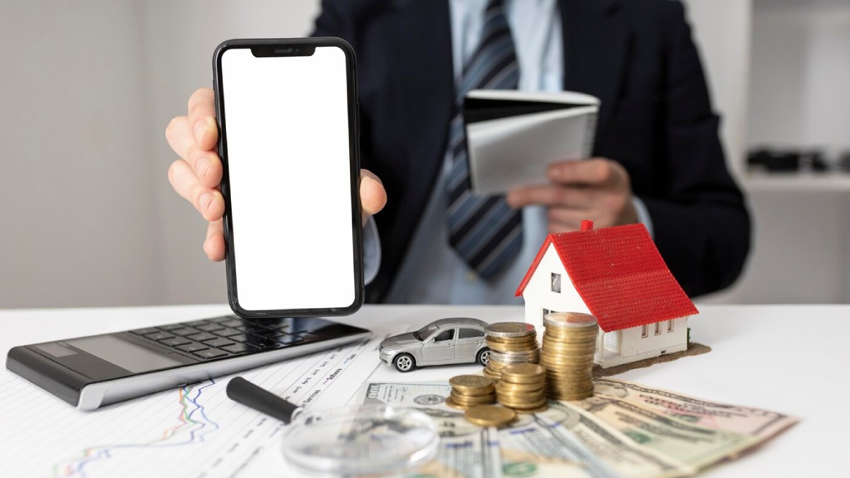 A businessman in a suit holds a smartphone, surrounded by money, a toy car, a model house, and a laptop, symbolizing finance and investments.
