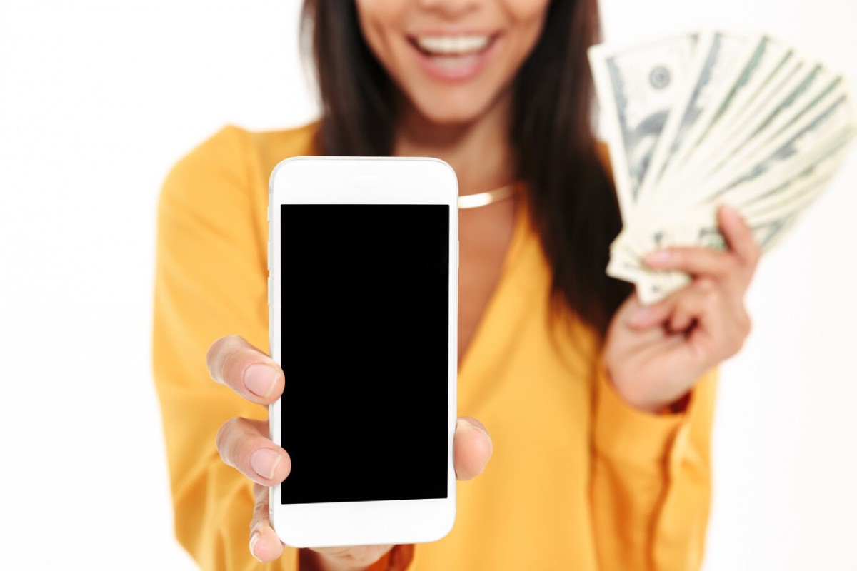 A smiling woman in a yellow top holds a blank smartphone in one hand and a fan of cash in the other, showcasing excitement.