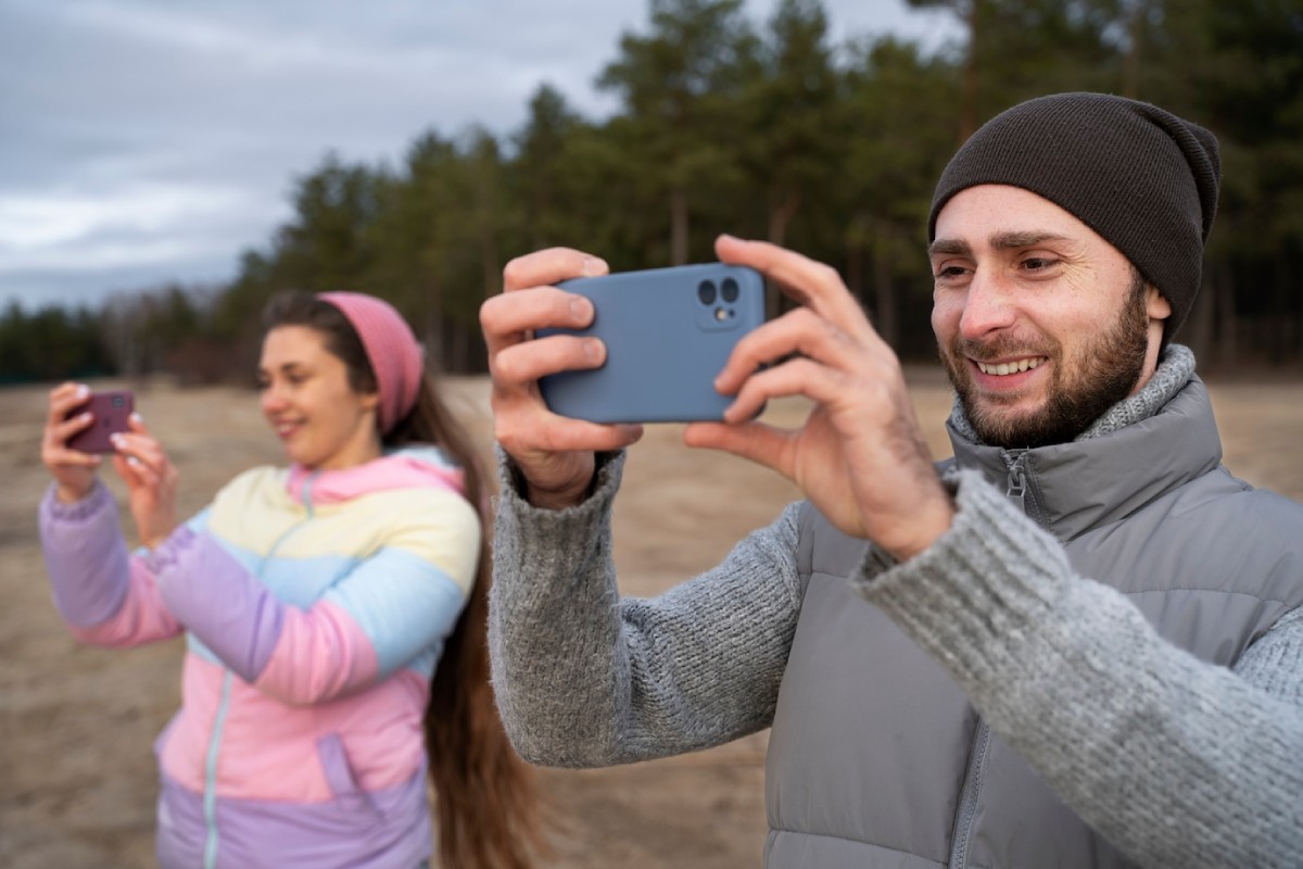 Two people are outdoors taking photos with their smartphones, surrounded by trees and a cloudy sky. One is in a colorful jacket, the other in a gray vest.