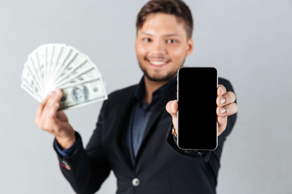 A person in a suit holds a smartphone in one hand and a fan of dollar bills in the other, against a gray background.