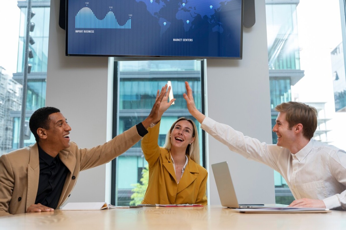 Three professionals enthusiastically engage in a high-five during a meeting, with a large screen displaying graphs and a world map behind them.