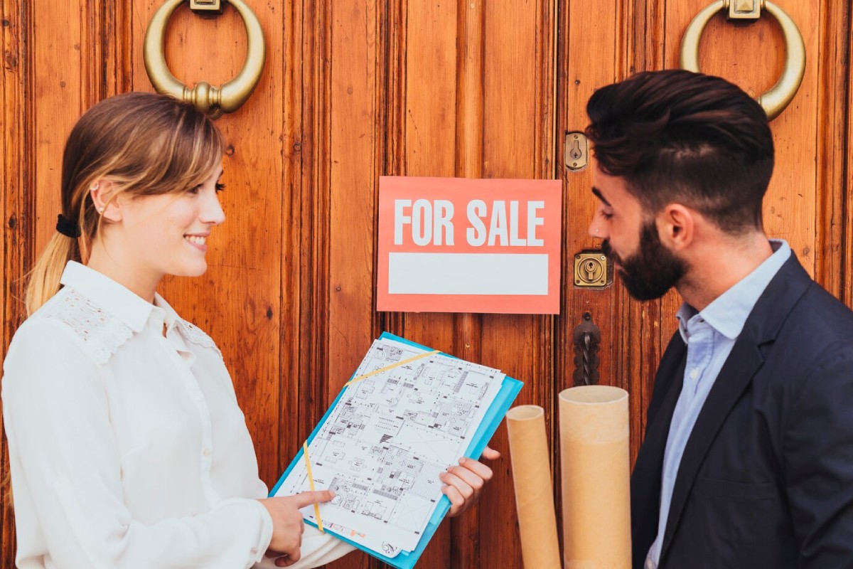 A woman in a white shirt holds a property plan while discussing a 'For Sale' sign on a wooden door with a man in a suit.