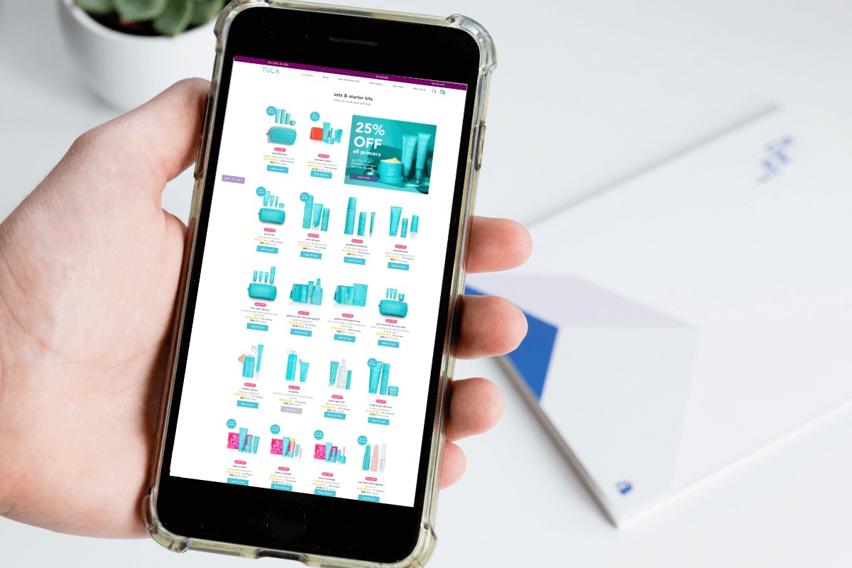 A hand holds a smartphone displaying Tula's website, showcasing skincare products with a 25% discount message prominently featured.