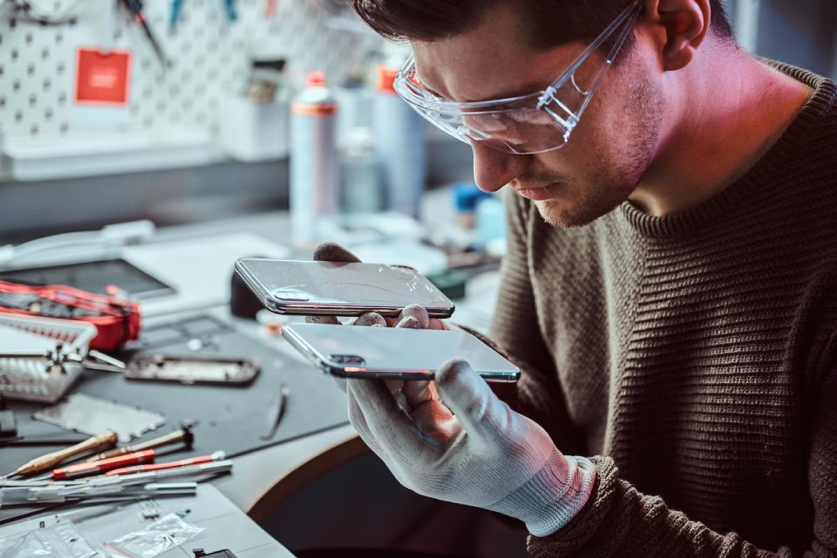 A person in a workshop examines two smartphone screens, one cracked and one intact, wearing gloves and surrounded by tools.