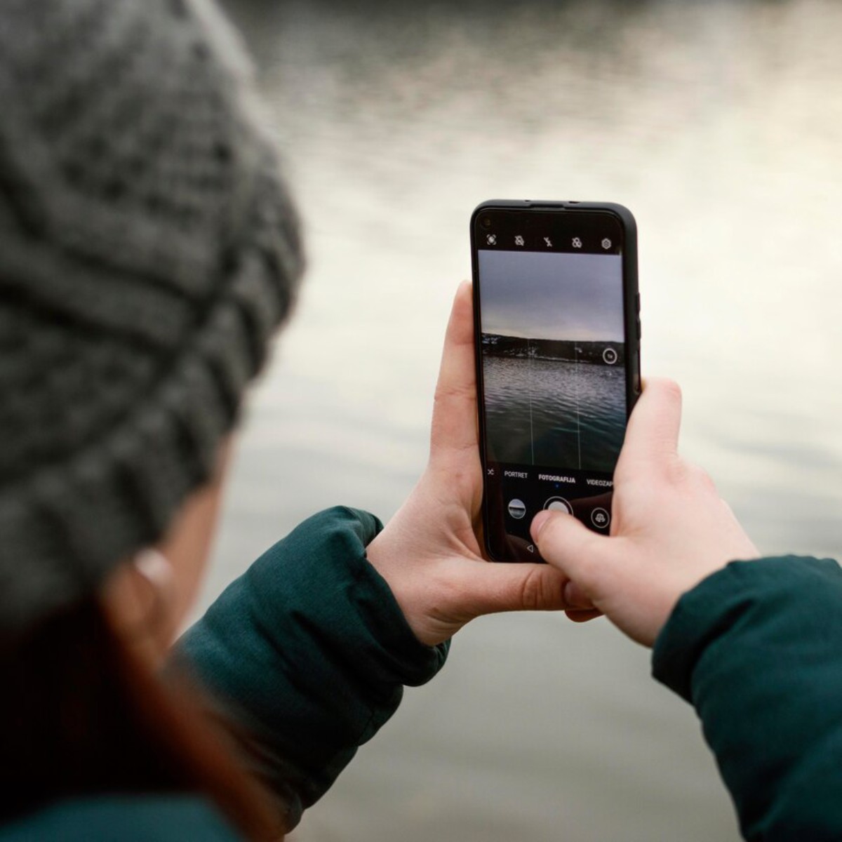 A person wearing a knit hat captures the serene water landscape with their smartphone, focusing on the camera screen.