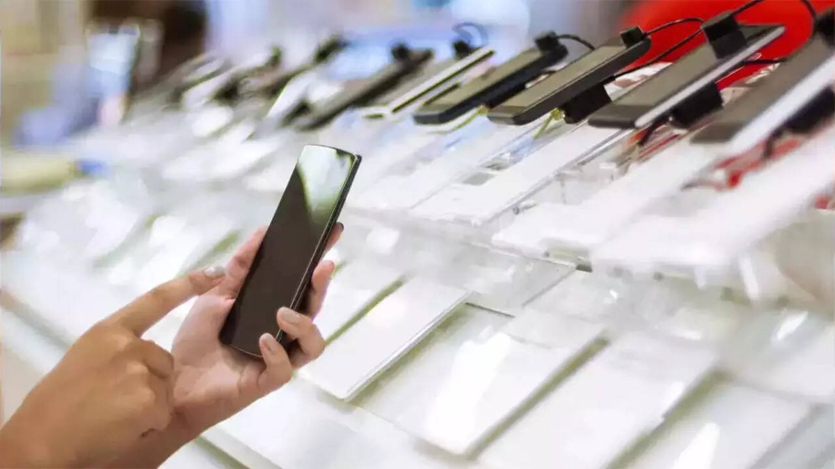 A hand holds a smartphone while browsing various displayed smartphones in a retail store.