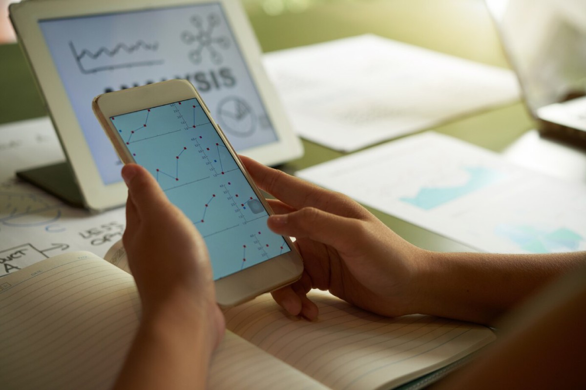 A person holds a smartphone displaying graph data, with a tablet and papers with charts on a desk, indicating data analysis work.