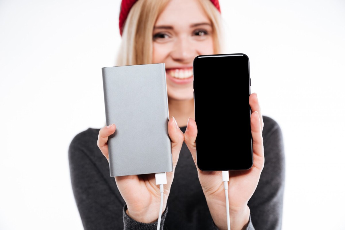 A person holds a silver portable charger in one hand and a black smartphone in the other, both connected with white charging cables.