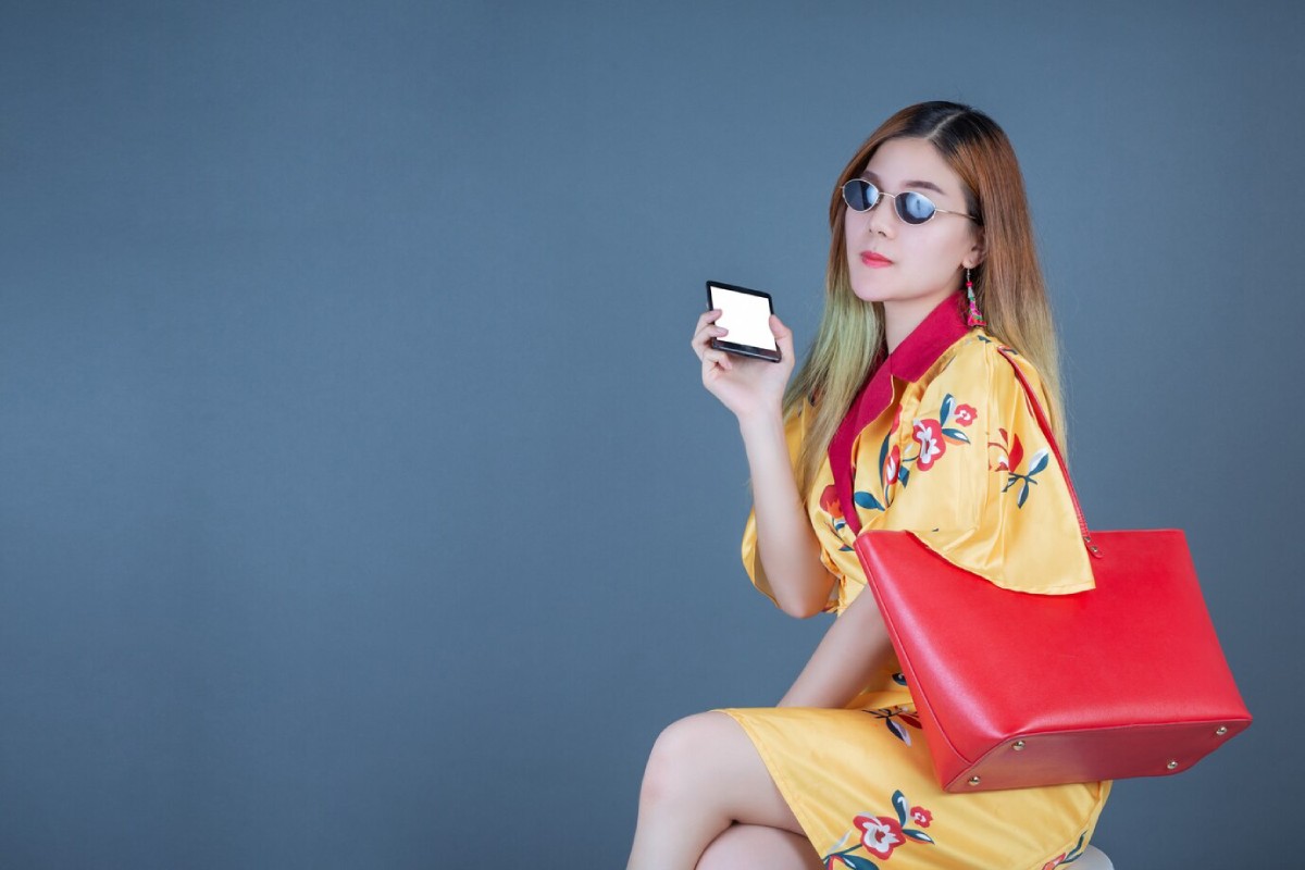 A woman in a floral yellow dress holds a small mirror while sitting, with a vibrant red handbag resting by her side against a blue backdrop.