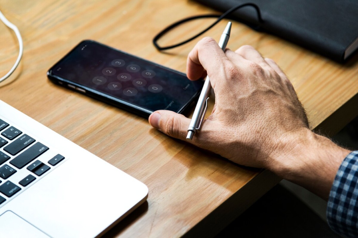 A person's hand holding a pen enters a passcode on a smartphone beside a laptop on a wooden desk.