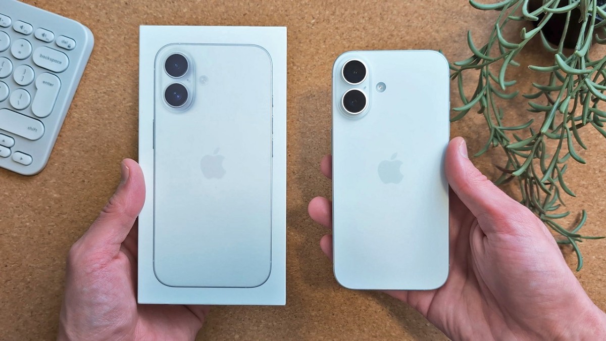 A person holds a new iPhone next to its white box, with a minimal keyboard and a green plant in the background.