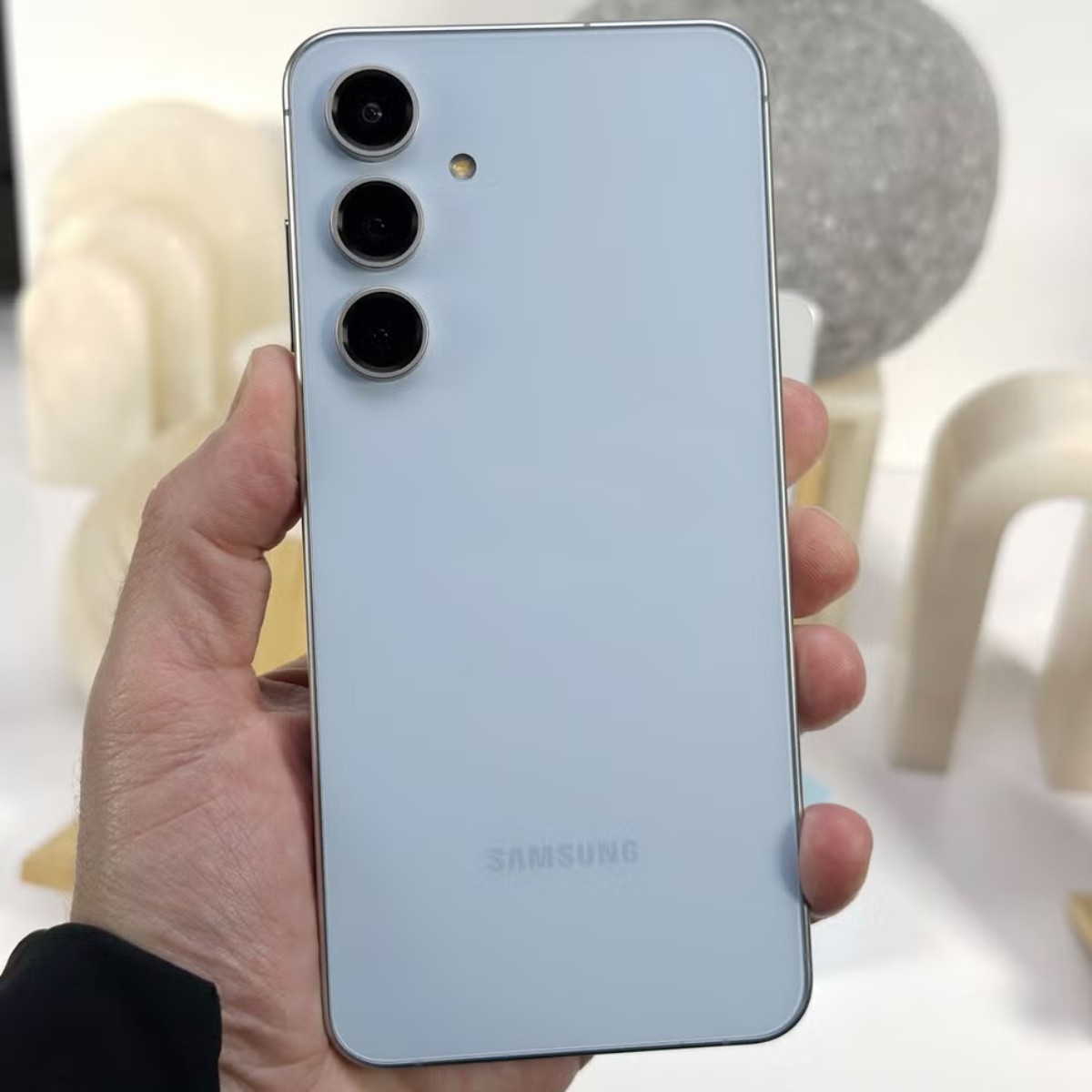 A person holds a light blue Samsung smartphone featuring three rear cameras, displayed against artistic sculptures in the background.