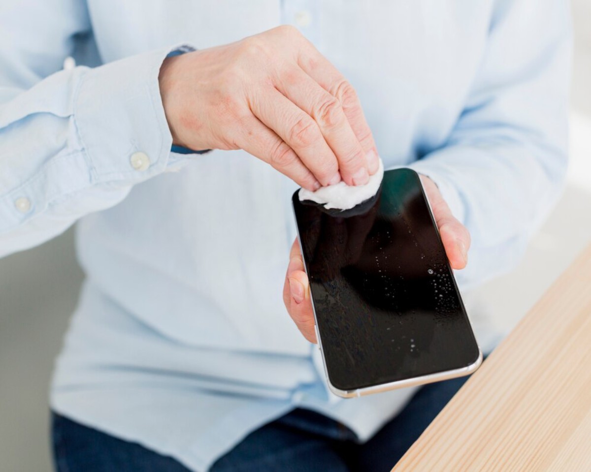 A person in a light blue shirt cleans a smartphone screen with a cotton pad, removing smudges and dirt.