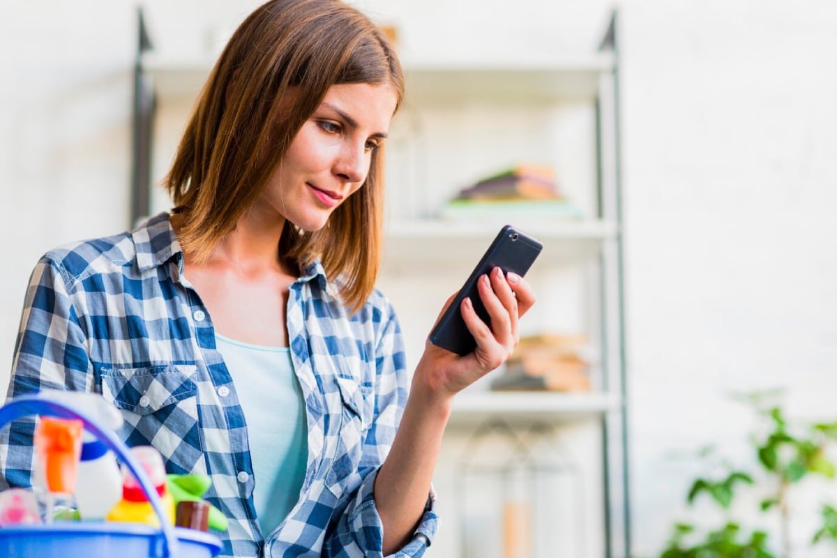 A woman in a checkered shirt holds a smartphone, surrounded by colorful cleaning supplies and a bright indoor space.