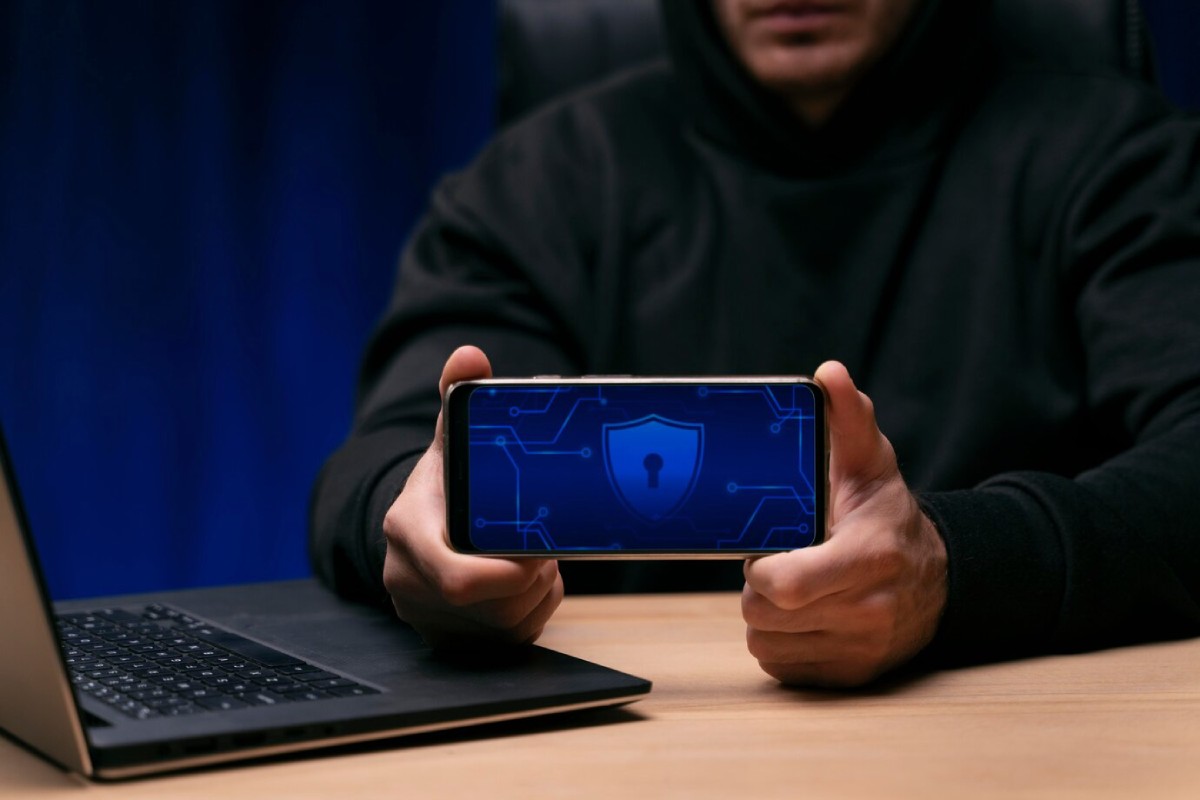 A person in a black hoodie holds a smartphone displaying a blue digital shield with a lock, next to a laptop on a wooden desk.
