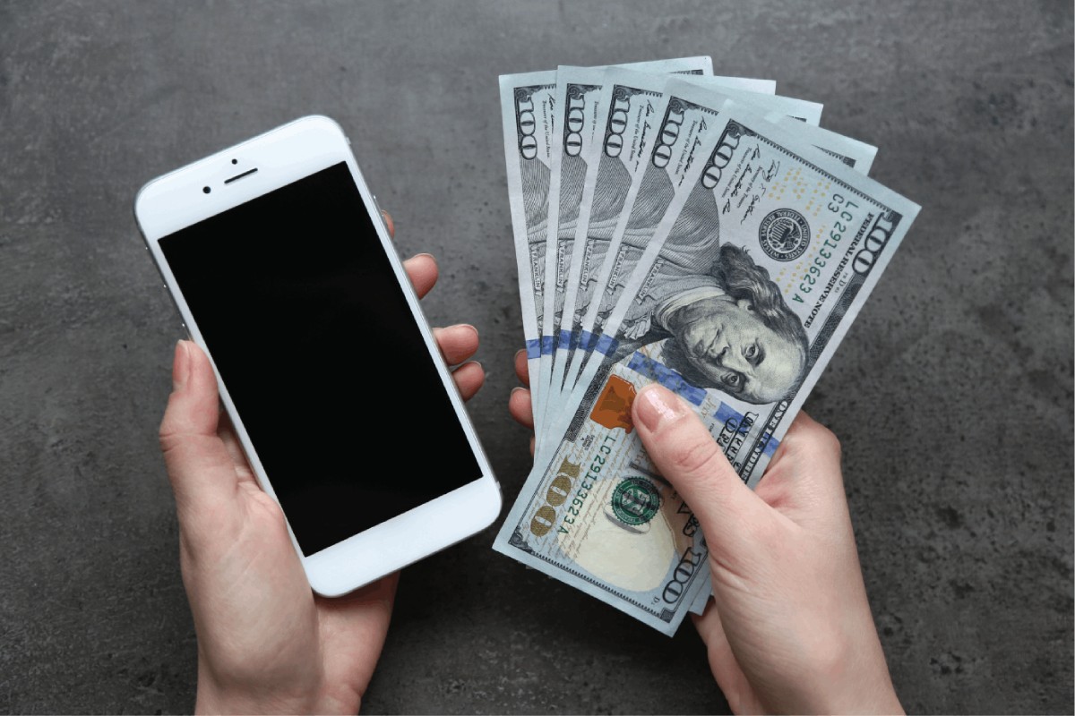 A person holds a smartphone in one hand and a bundle of hundred-dollar bills in the other, against a gray background.