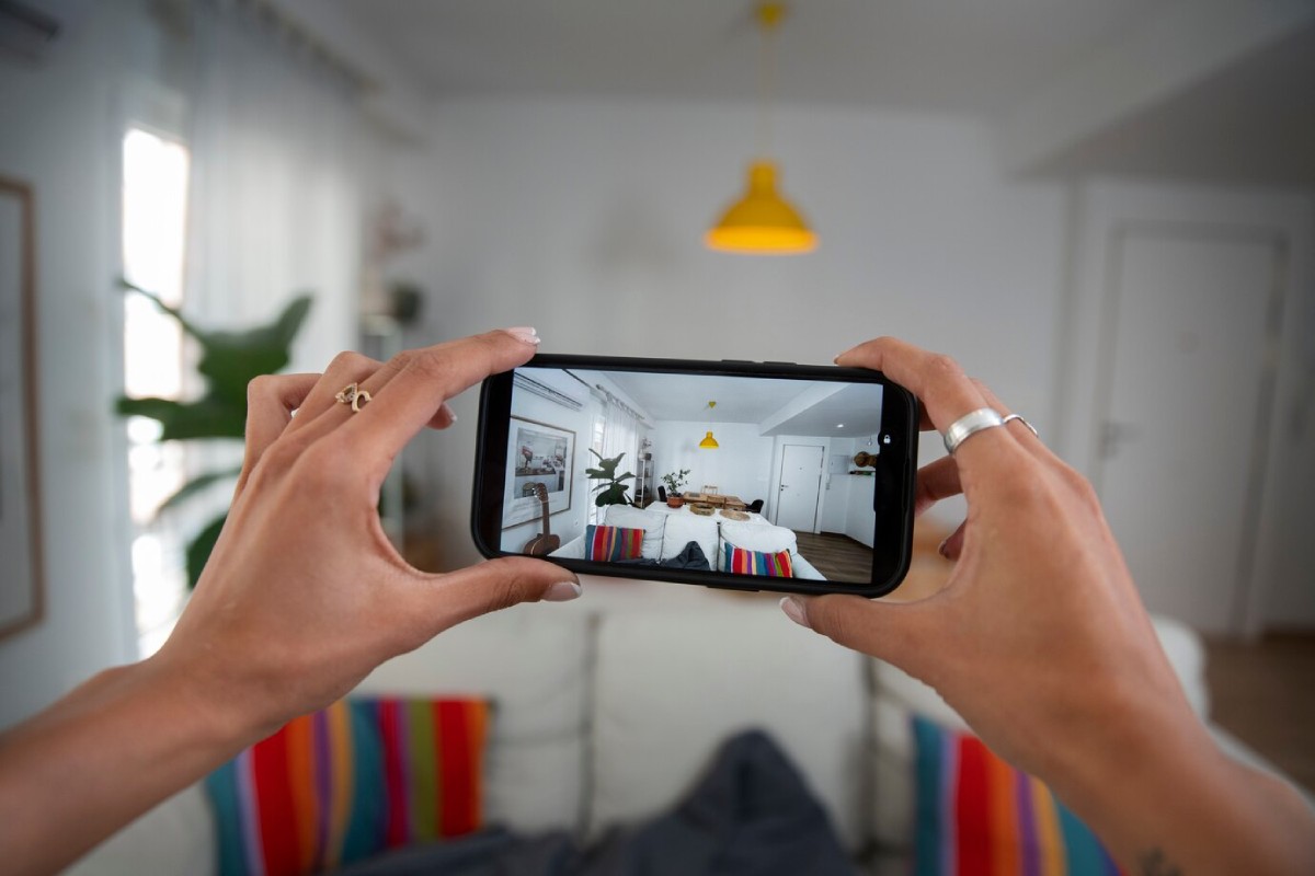 A person takes a photo of a cozy living room with a vibrant sofa and a yellow pendant light, using a smartphone.