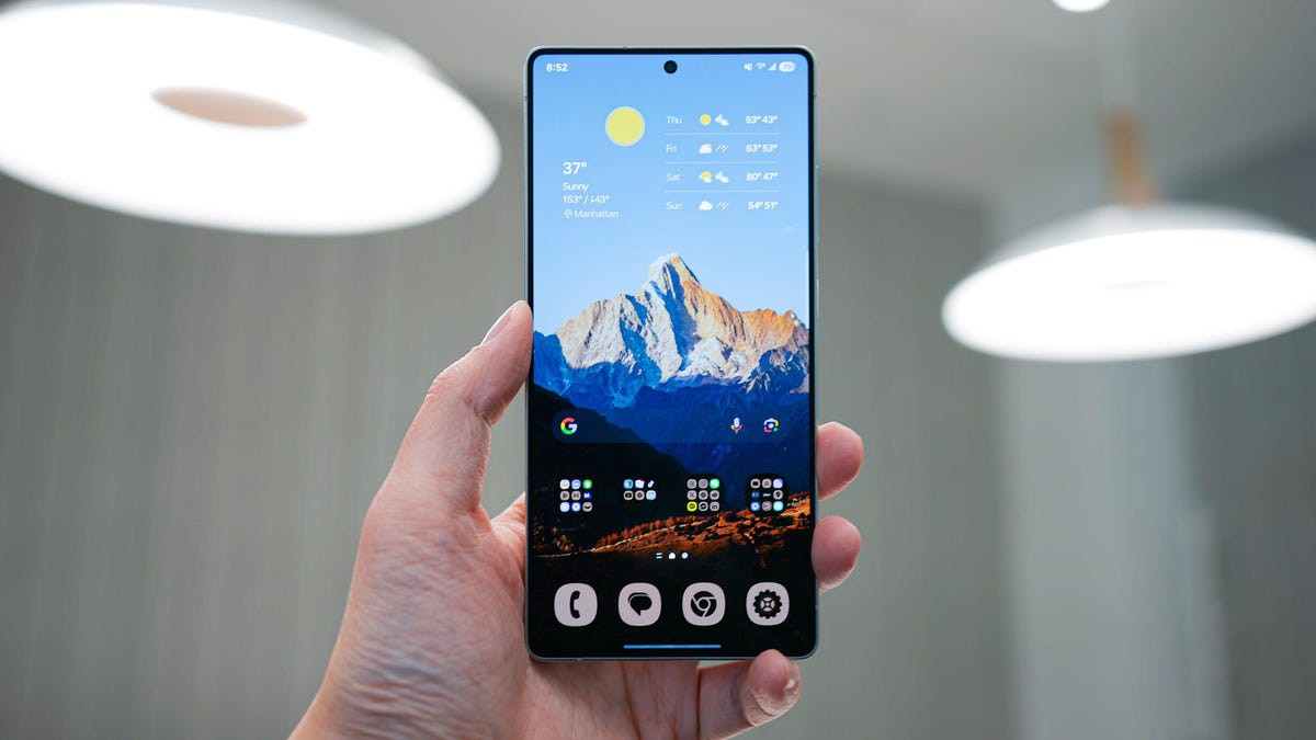 A hand holds a smartphone displaying a mountain landscape and weather widget, with app icons below and soft ceiling lights in the background.
