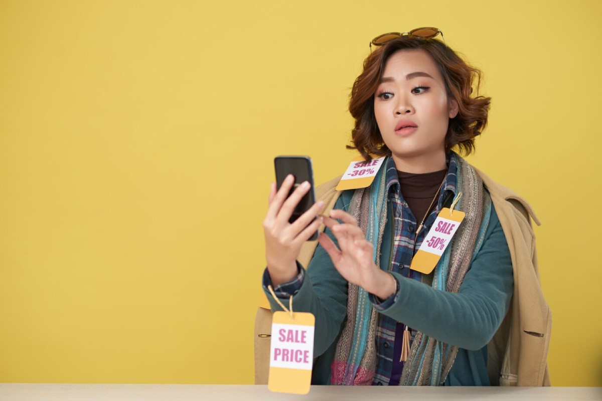 A person with curly hair, holding a smartphone, surrounded by various sale tags, against a bright yellow background.