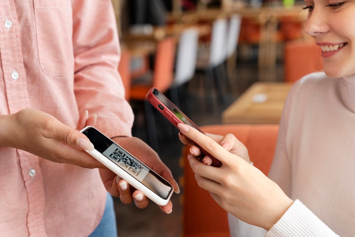 A person in a pink shirt holds a white phone displaying a QR code, while another person scans it with a red phone at a cafe.